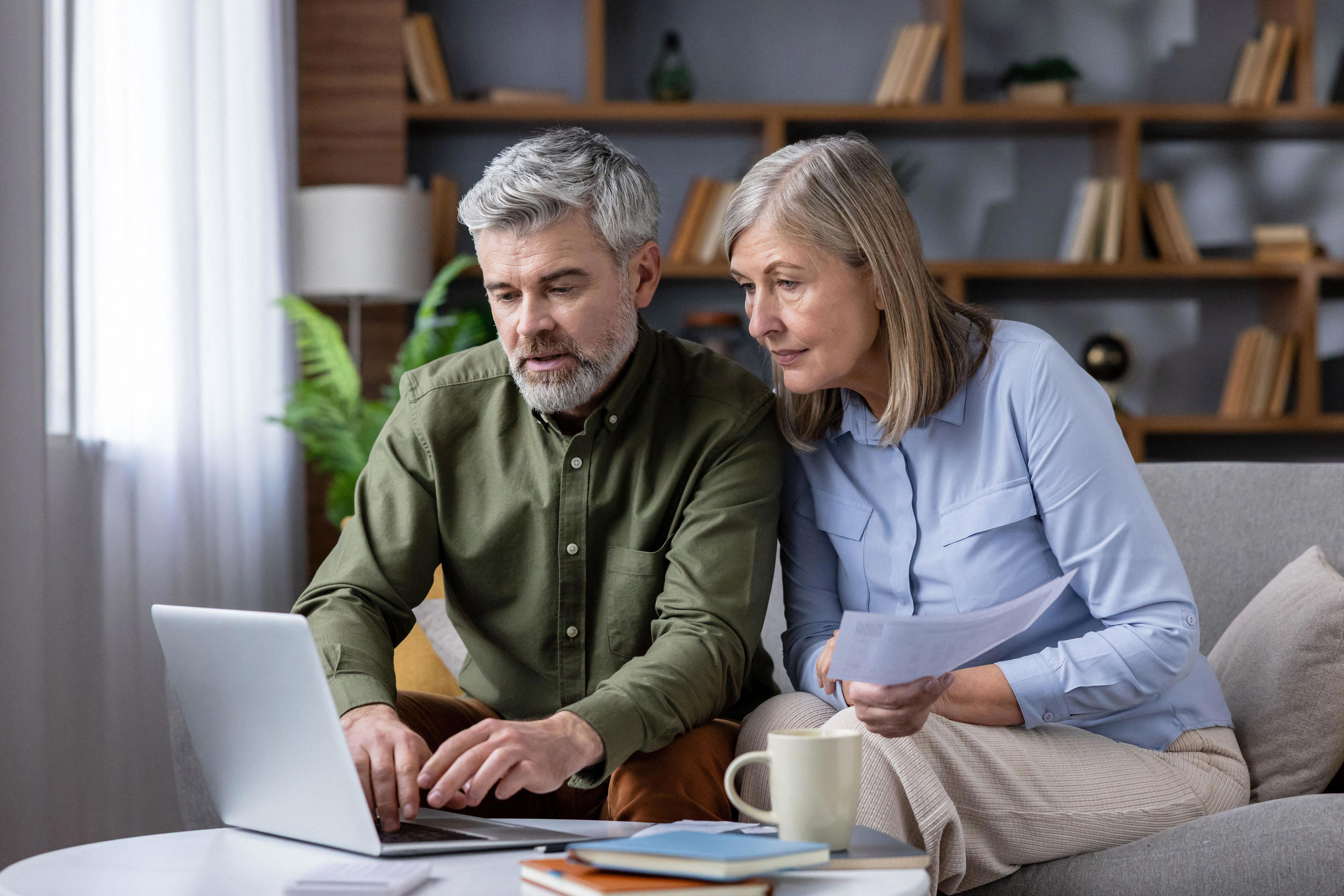 3DT2T89 Mature couple on sofa in cozy living room using laptop and papers to review bills, budget and retirement plans, focused on savings, mortgage and investments for future