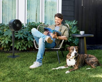Person relaxing outdoors with a dog by their side, reading. Nearby stands a fan labeled