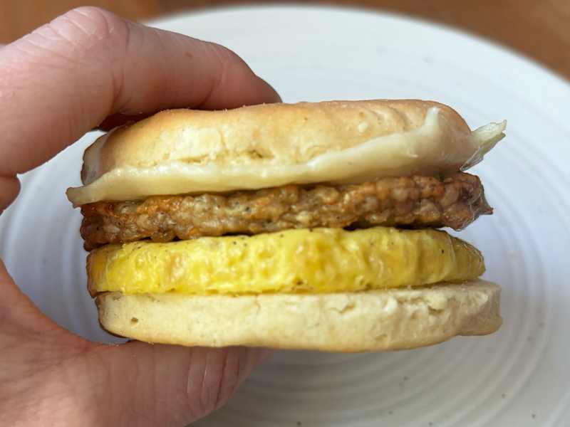 A hand holding a breakfast sandwich with a sausage patty, egg, and a soft bun on a white plate.