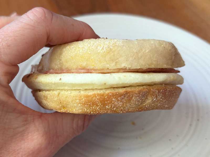 A hand holding a sandwich with a soft bun, cheese, and ham, placed on a white plate.