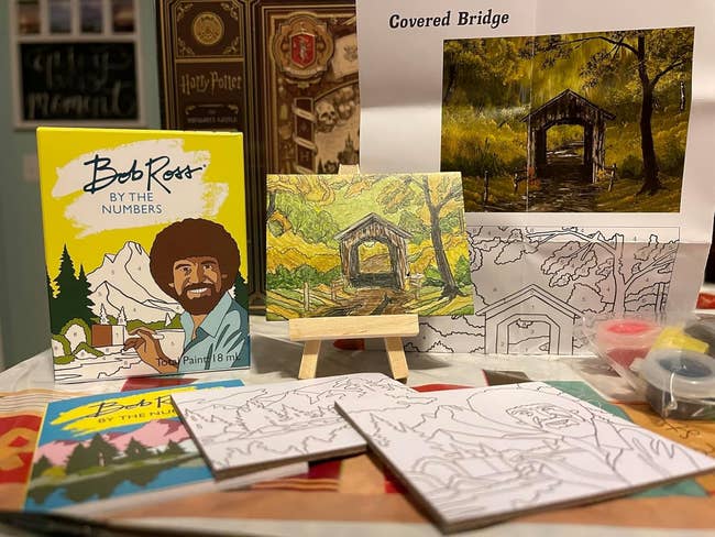 Bob Ross paint-by-numbers kit displayed on a table with canvas, paints, and a small easel featuring a covered bridge scene