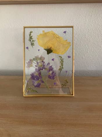 Pressed flowers in a gold frame, featuring a large yellow flower and smaller purple blooms, displayed on a wooden surface