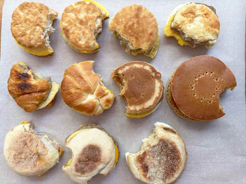 Assorted breakfast sandwiches with bites taken out, featuring biscuits, croissants, and pancakes on parchment paper.