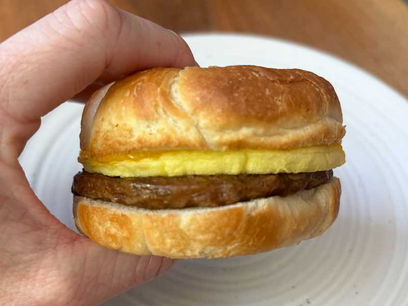 A hand holding a breakfast sandwich with a flaky croissant, egg, and sausage patty on a white plate.