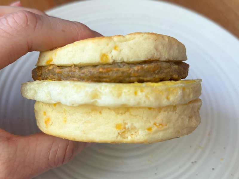 A hand holding a biscuit sandwich with sausage and egg, placed on a white plate.
