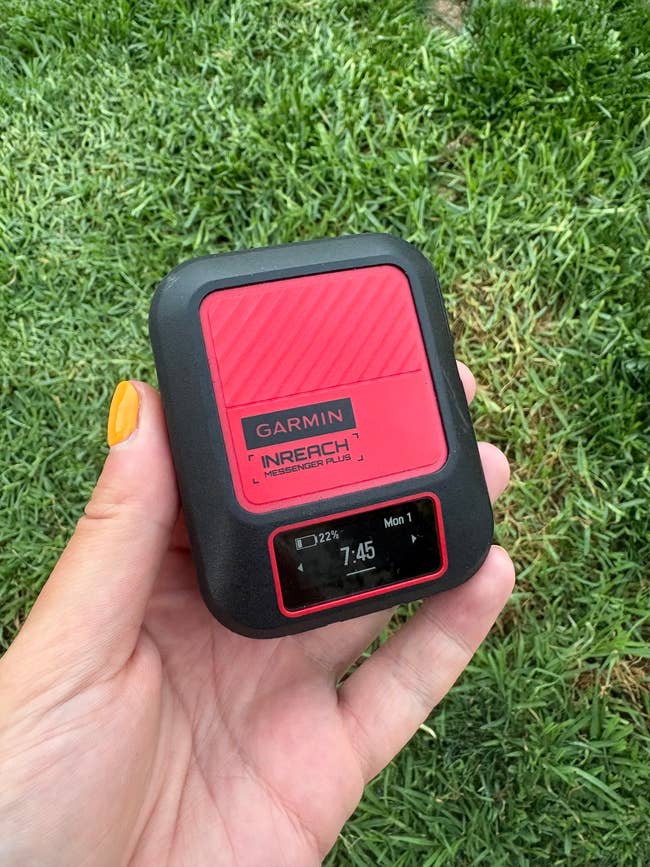 Hand holding a Garmin inReach Mini satellite communicator, showing a digital screen with time and date. Lawn background