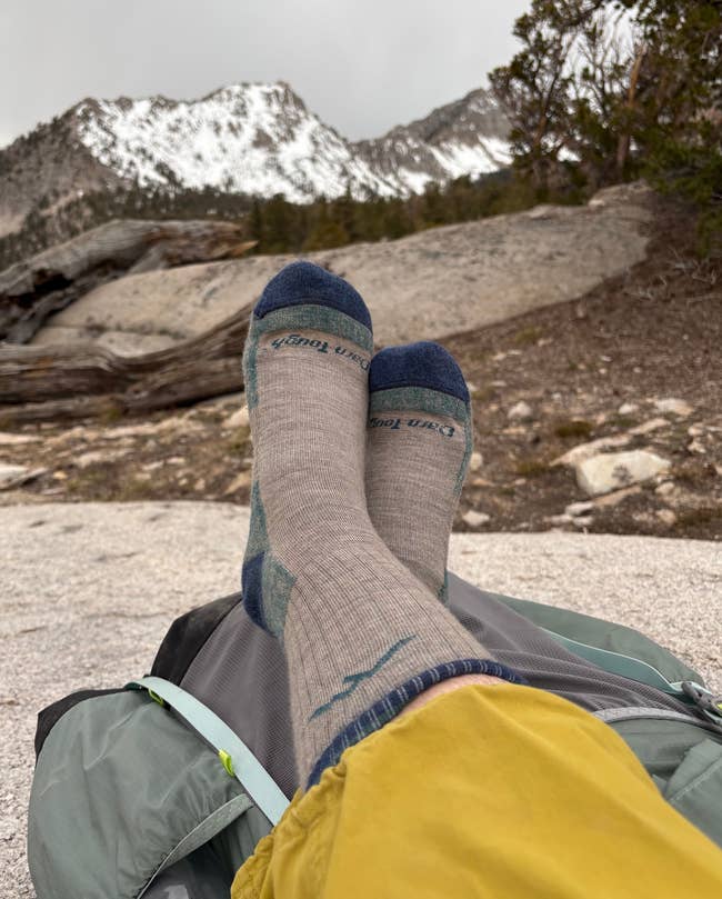 Person relaxing outdoors with feet in gray hiking socks, resting on a backpack. Snow-capped mountains and trees are visible in the background