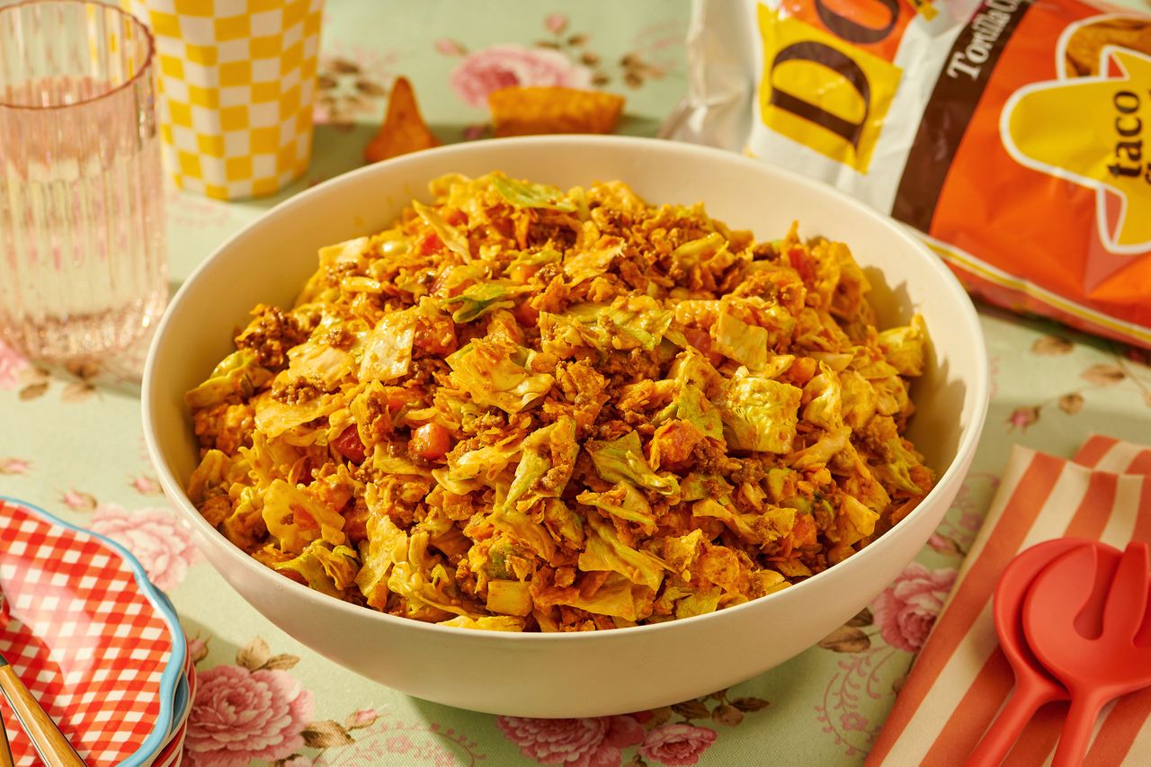 A bowl of taco salad with lettuce cheese and crumbled Doritos placed on a decorated table with a bag of Doritos nearby