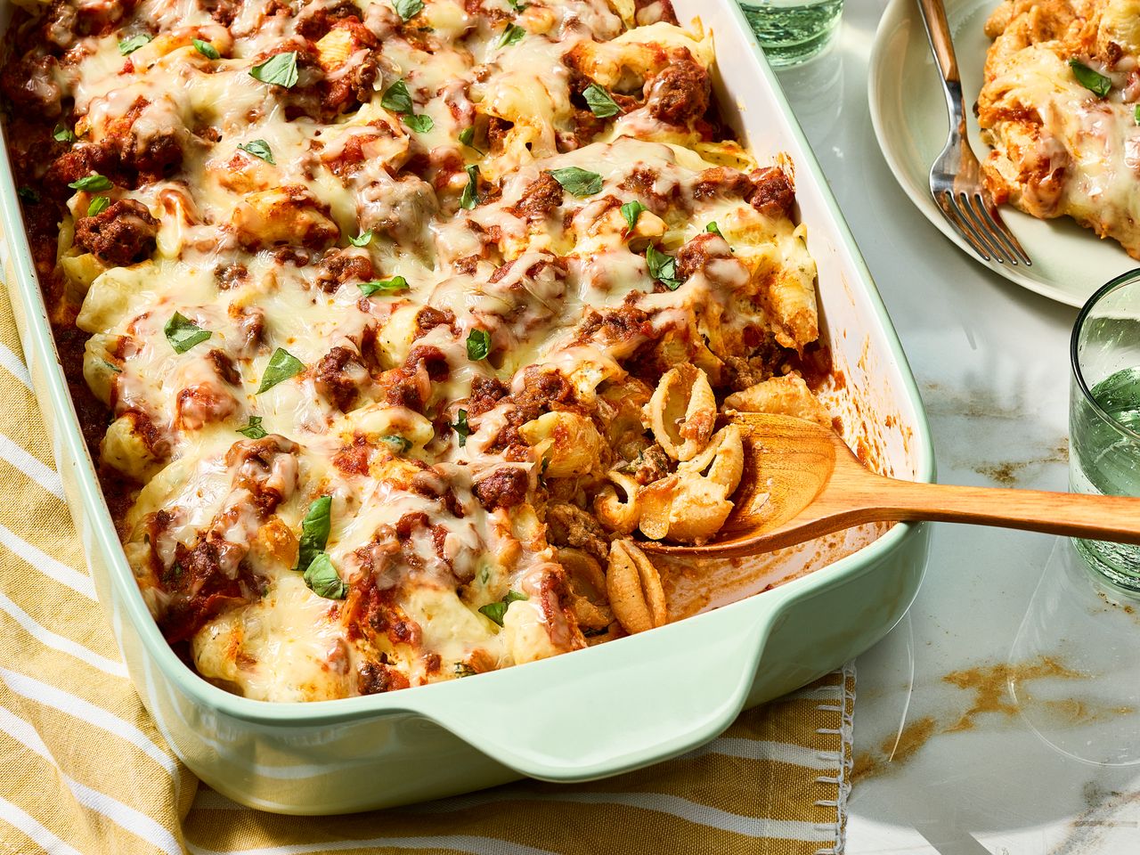 Unstuffed Shells Pasta Bake garnished with fresh basil in a baking dish with a wooden spoon, next to a plate and a glass