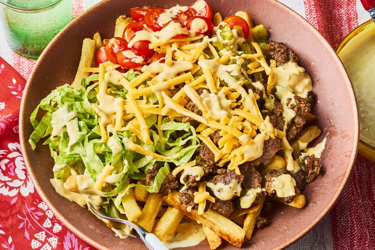 A cheeseburgerinspired salad bowl with ground beef shredded cheese lettuce tomatoes and a sauce over fries in a bowl