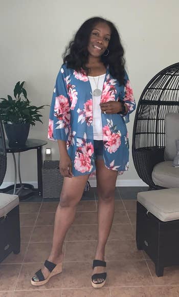 Person smiling and posing indoors, wearing a floral kimono-style outfit with platform sandals. Cozy seating area with pillows in the background