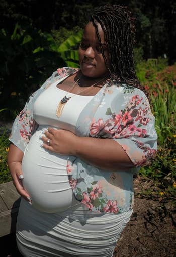 Pregnant woman in a fitted dress and floral shawl stands outdoors, gently holding her belly