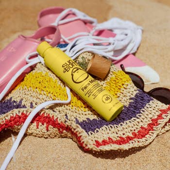 Sunscreen bottle on a woven beach bag with sunglasses and sandals beside it on sand, suggesting essential beach gear