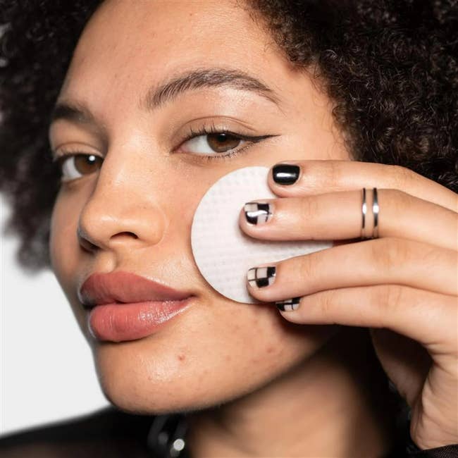 Person applying skincare product with a circular pad to their cheek. They have rings on their finger and manicured nails with a geometric black design