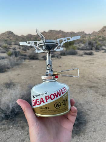 A portable camping stove is held in a hand outdoors attached to a fuel canister