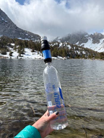 Hand holding a water bottle with a filter attached, with a scenic mountain lake and snowy peaks in the background
