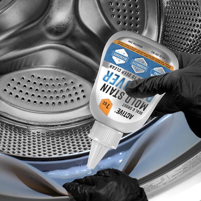 Gloved hands hold a mold and stain remover bottle, pouring it into an open washing machine