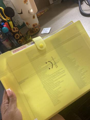 Hand holding a translucent yellow folder with a smiley face design, containing organized documents. Desk supplies visible in the background