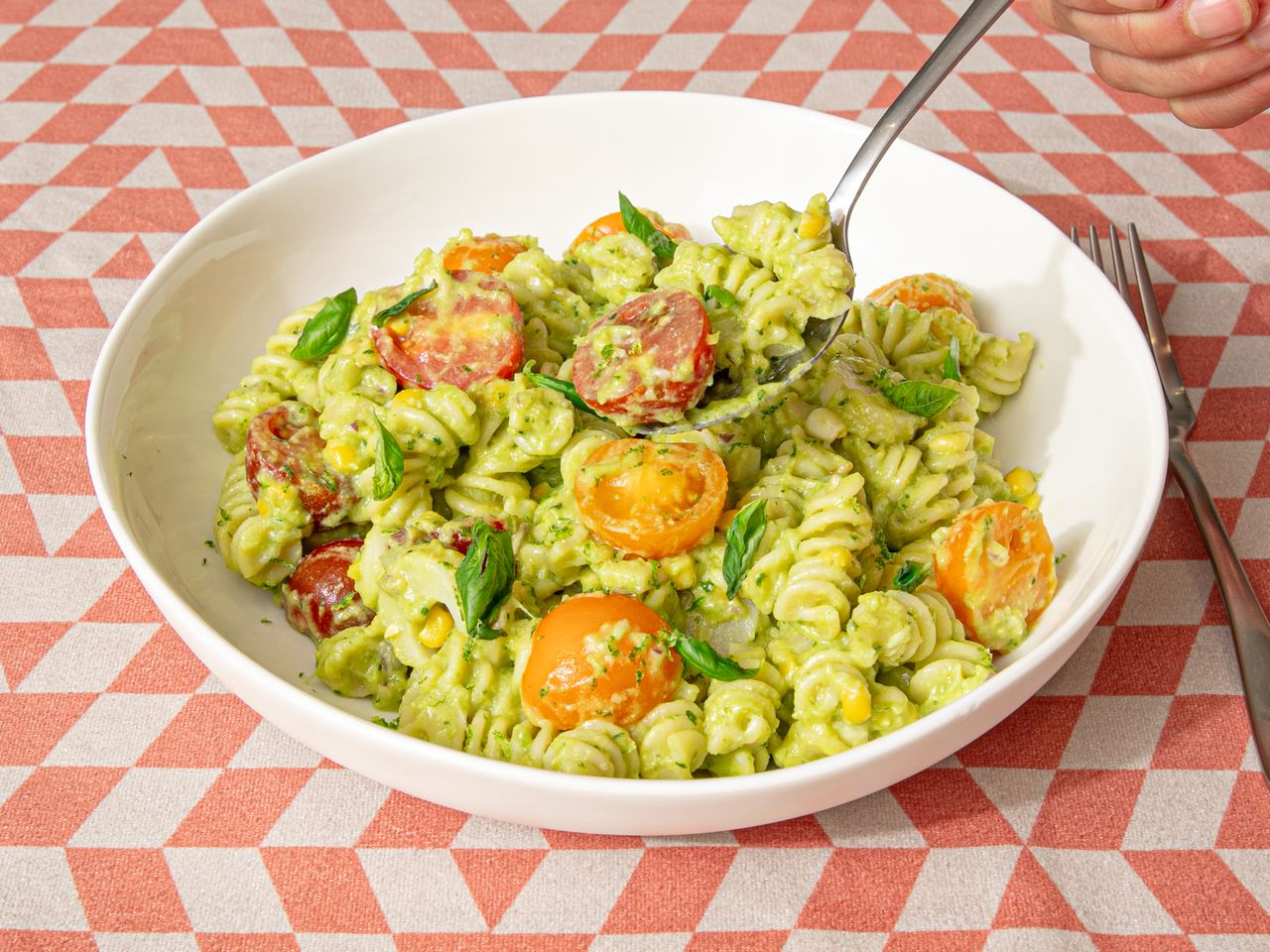 Bowl of pasta with pesto and cherry tomatoes on a patterned tablecloth hand holding a fork