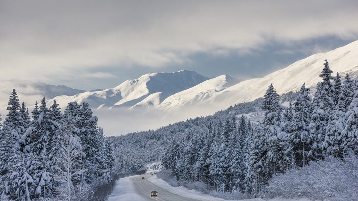A car drives down the highway surrounded by snow-covered trees with mountains in the distance.