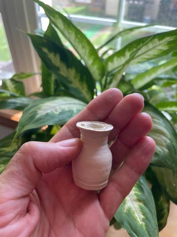 A hand holds a small, handcrafted ceramic vase against a backdrop of lush indoor plants
