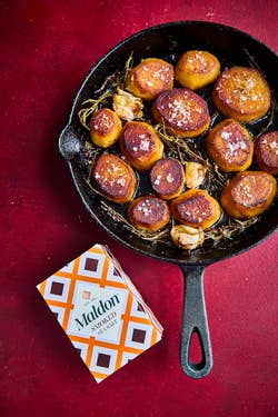 Roasted potatoes with garlic and rosemary in a skillet, topped with salt flakes. A package of Maldon Smoked Sea Salt is nearby
