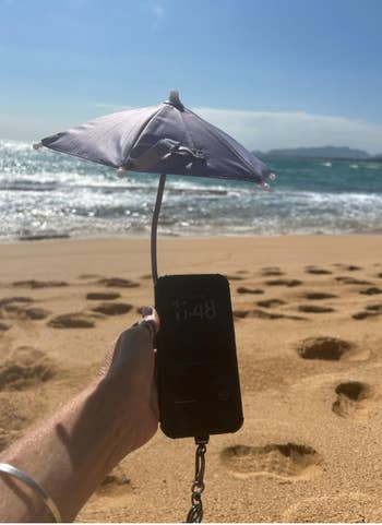 Phone held on sandy beach under tiny umbrella, displaying time 11:48 AM. Waves in background. Novelty phone accessory for sun protection