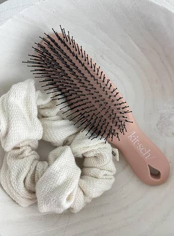 Hairbrush and two soft fabric hair scrunchies placed in a decorative bowl, featured in a shopping article