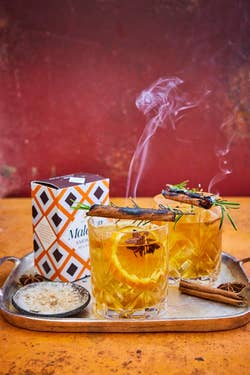 Two glasses of smoked cocktails with herbs and orange slices on a tray, beside a spiced rum box, suggest a cozy beverage experience