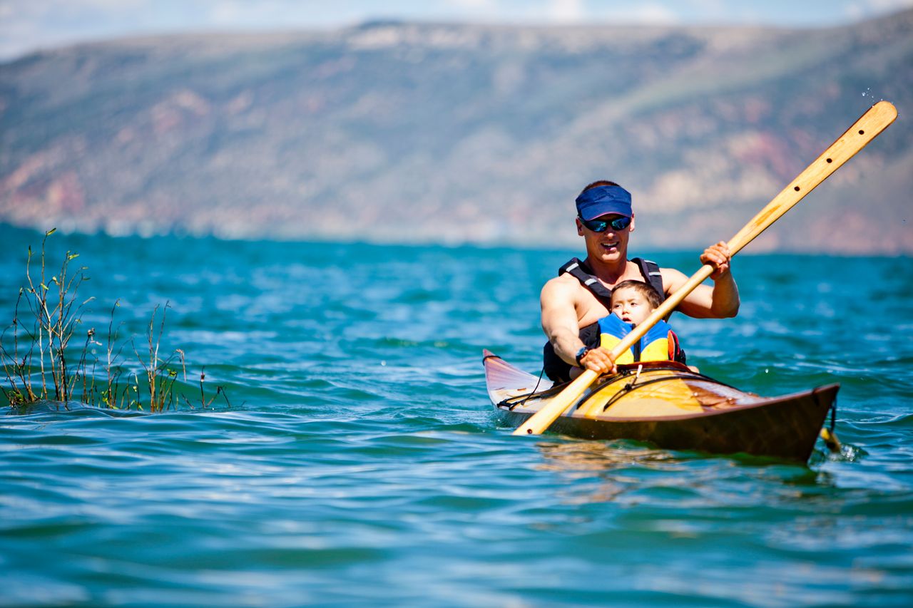 A man and a child kayaking together on a lake with mountains in the background wearing life jackets and paddling