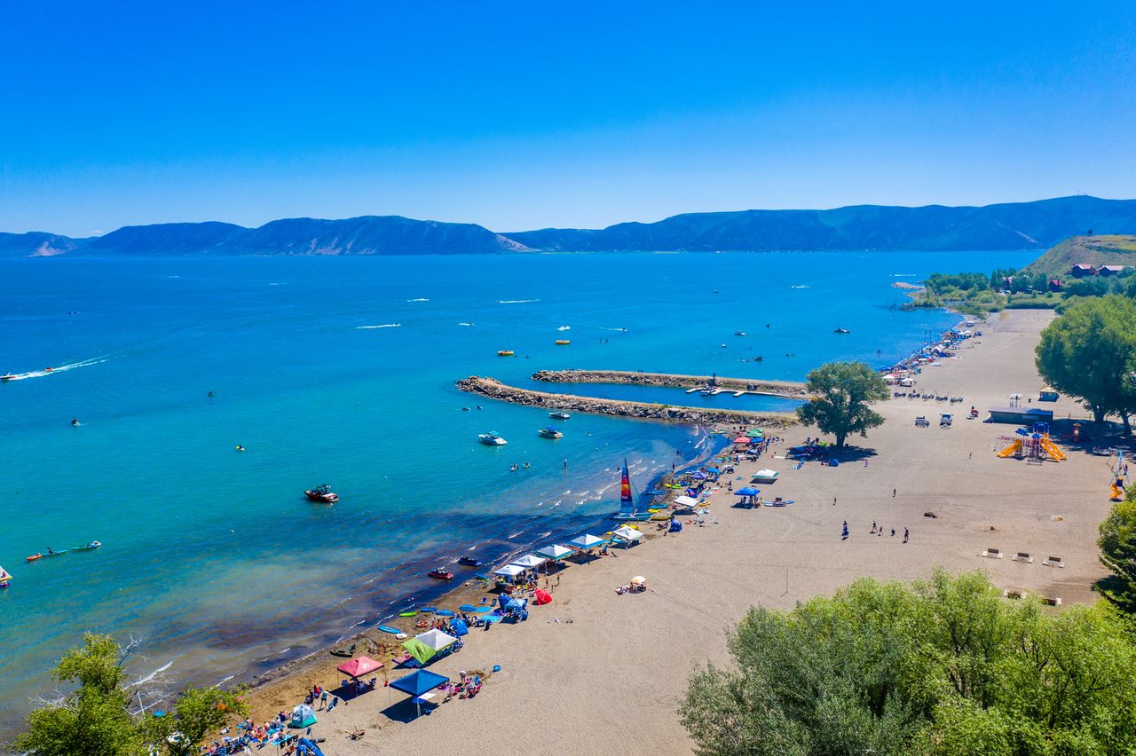 A scenic beach area with people enjoying recreational activities by a lake with distant mountains across the water