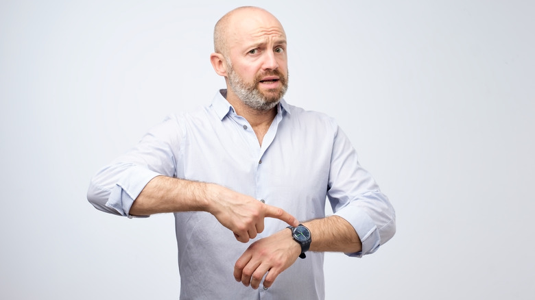 A man wearing a white button-down shirt pointing at his wristwatch with a concerned expression.