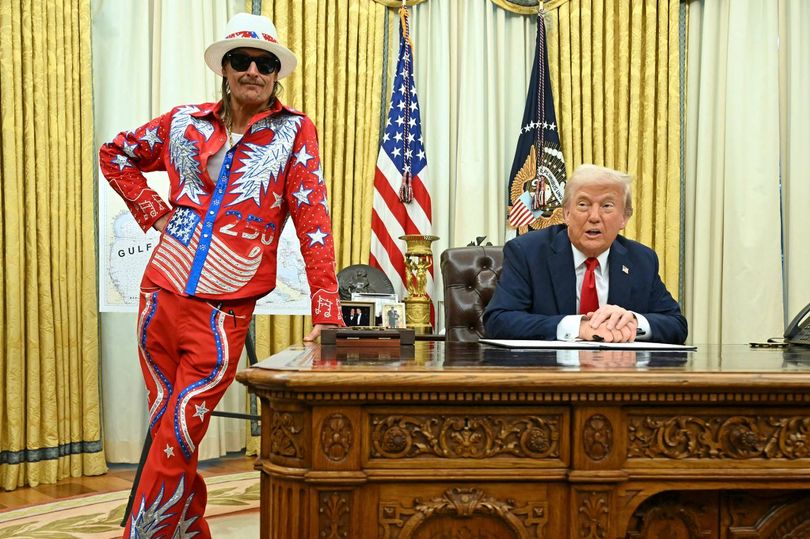 An individual dressed in a colorful, patriotic-themed costume stands behind a desk, facing an individual seated in a chair, who appears to be engaged in an official activity. Behind them, two American flags are displayed against a backdrop of yellow drapes.