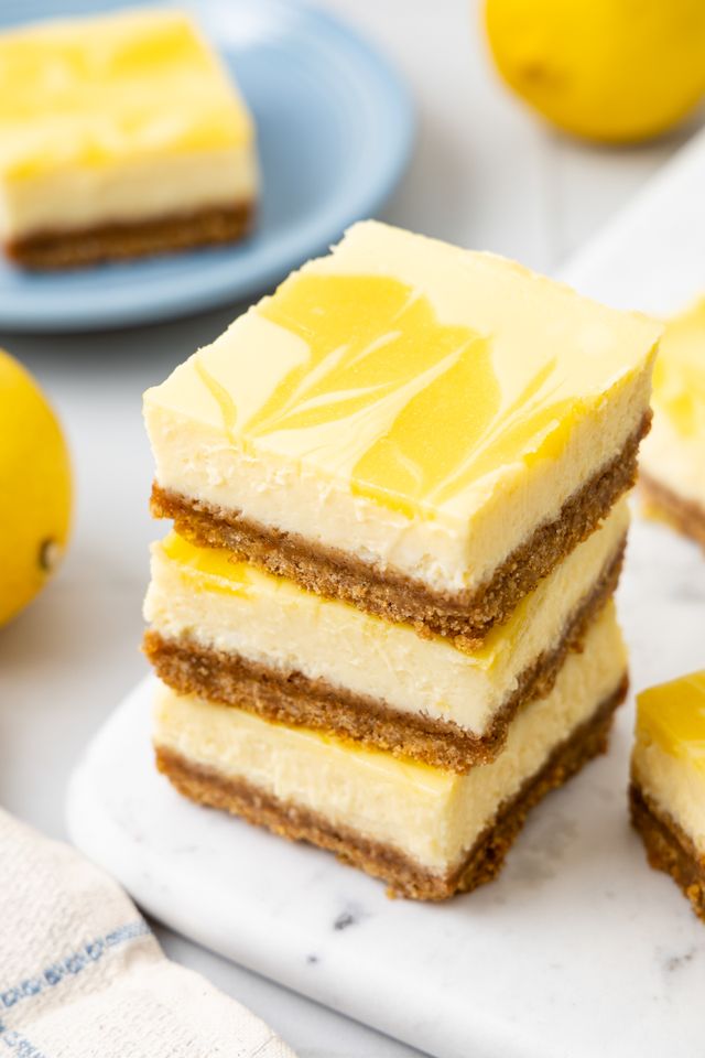 Stack of Lemon Cheesecake Bars on a Marble Tray Next to More Bars, and in the Surroundings, a Kitchen Towel, Lemons, and Another Bar on a Plate