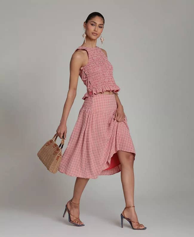 Person in a stylish checked two-piece ensemble with a halter top and flared skirt, holding a woven handbag and wearing strappy heels