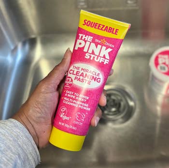 Person holds tube of The Pink Stuff cleaning paste above a sink. The tube claims it's easy to rinse and effective for multi-purpose cleaning