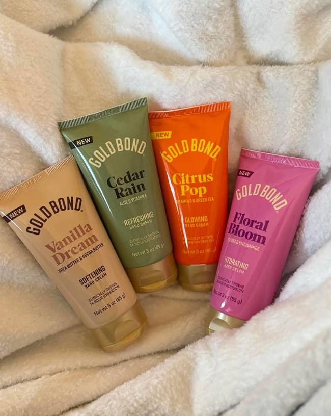 Four tubes of Gold Bond hand cream rest on a soft blanket: Vanilla Dream, Cedar Rain, Citrus Pop, and Floral Bloom, offering a variety of scents
