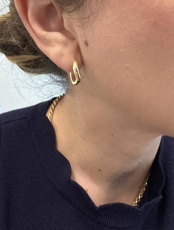 Close-up of a person wearing a gold hoop earring and a gold necklace, with a scalloped neckline visible