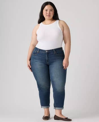 Person wearing a white tank top, blue jeans, and brown flats poses confidently against a plain background