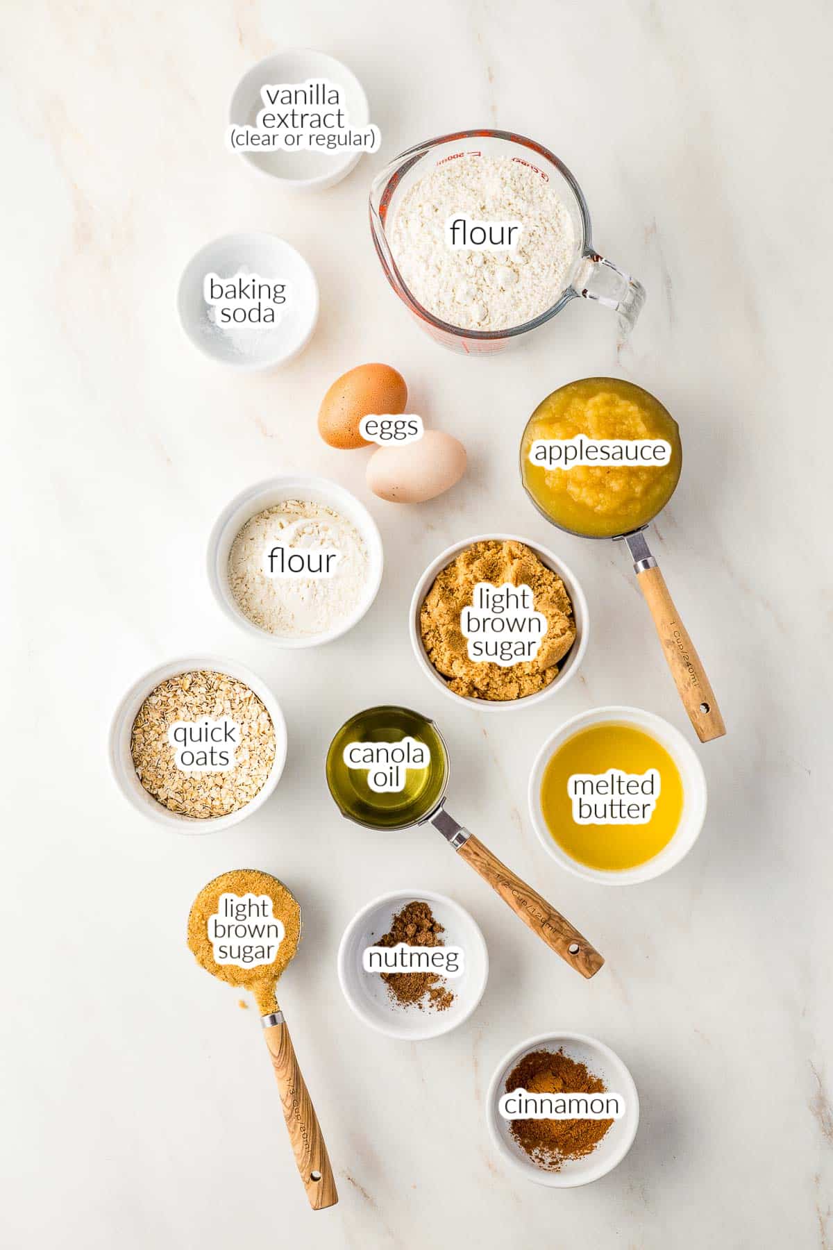 Baking ingredients for Applesauce Crumb Cake including flour, oats, eggs, applesauce, canola oil, melted butter, brown sugar, cinnamon, nutmeg, baking soda, and vanilla extract on a white surface.