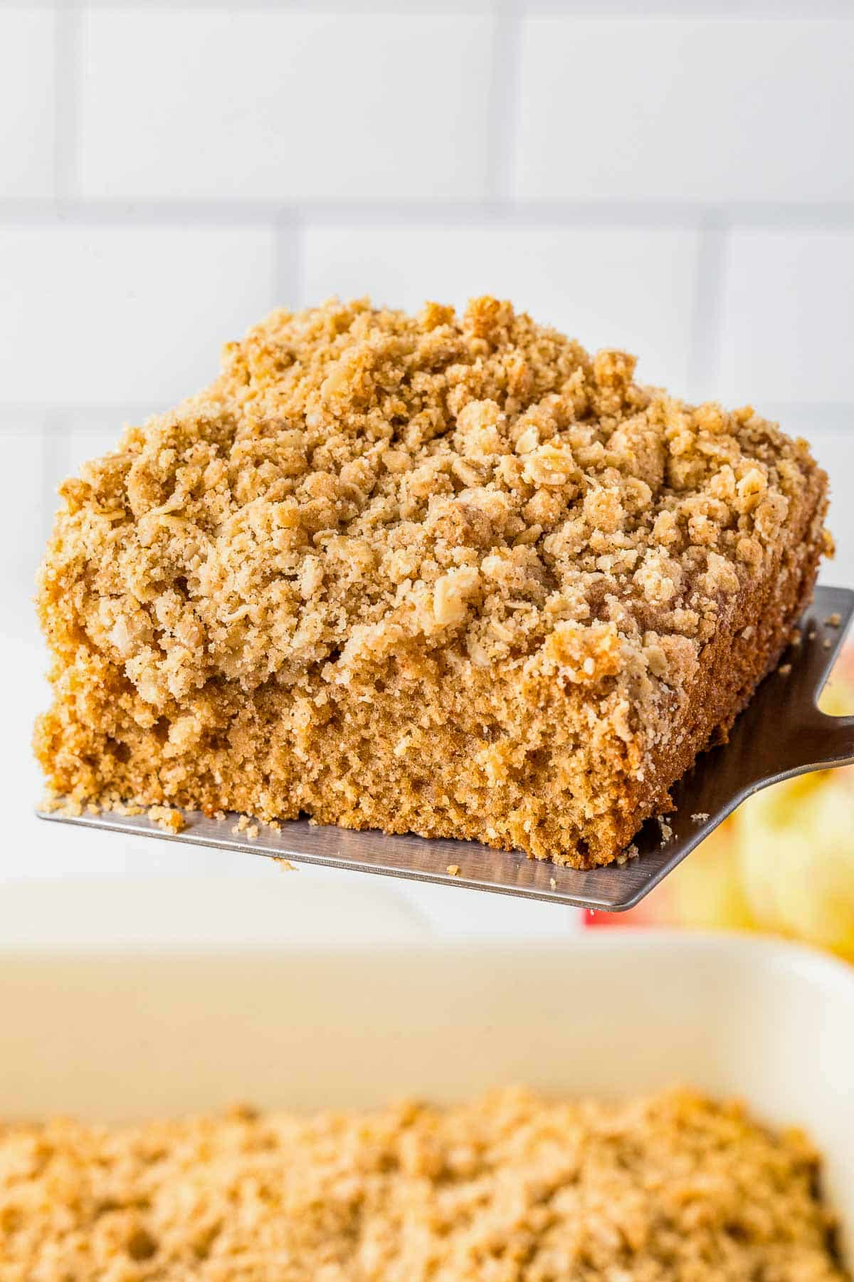 A close-up of a square slice of Applesauce Crumb Cake on a metal spatula, with a white tiled background.