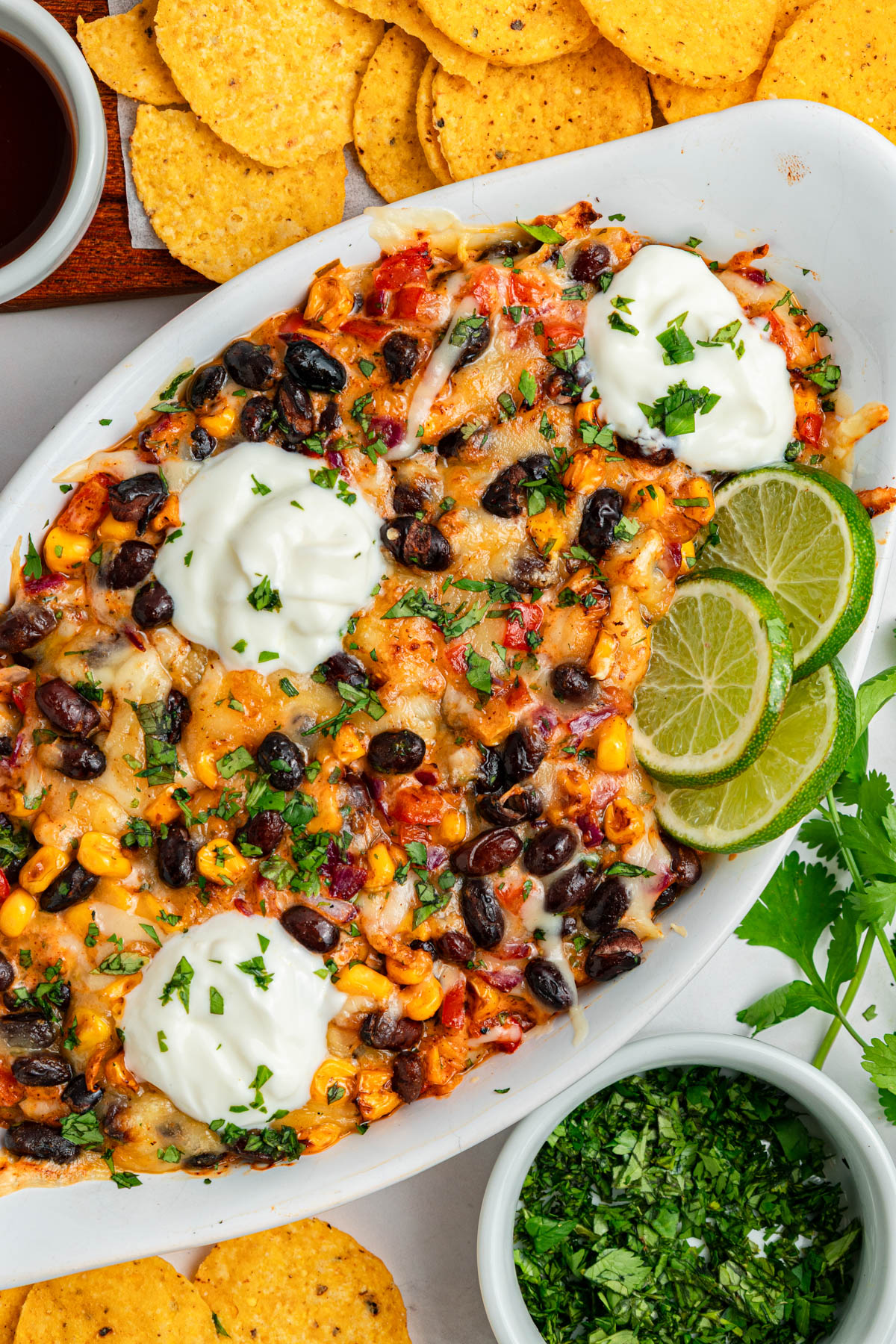 A baked dish topped with melted cheese, black beans, corn, and herbs, garnished with sour cream and lime slices, served with tortilla chips.