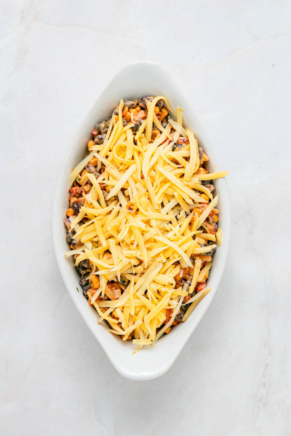 An oval white baking dish filled with a mixture of vegetables, topped with a layer of shredded cheese.