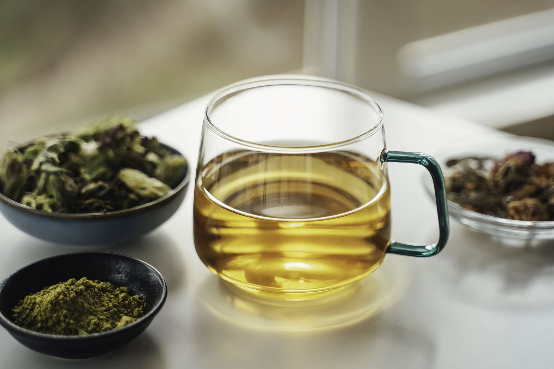 A glass cup of tea with three small dishes containing herbs and spices on a tabletop