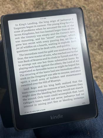 A person is reading a Kindle device displaying a page from a book about a king's challenges and strategies in a fictional realm