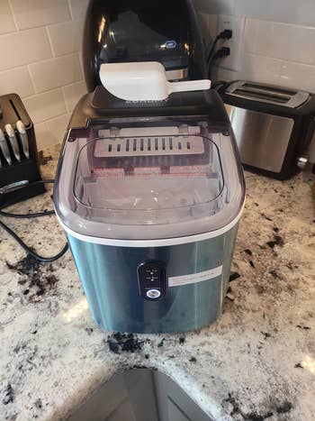 Home kitchen with a countertop ice maker, scoop on top, surrounded by appliances