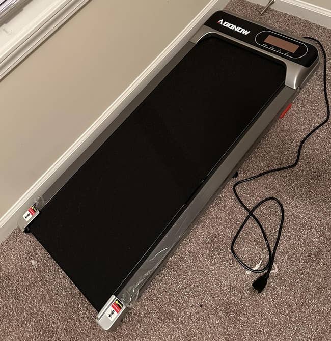 A foldable treadmill is placed in the corner of a carpeted room near a window with closed blinds