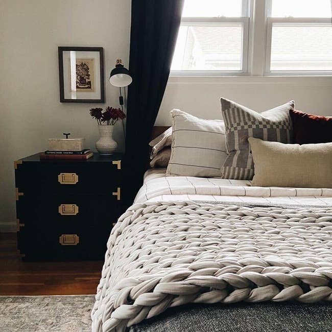 Cozy bedroom with a knitted blanket on the bed, decorative pillows, a nightstand with books, a lamp, and framed wall art