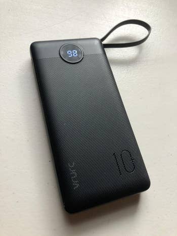 Portable power bank with a LED display showing 98% charge. Sleek design with a subtle pattern and a strap for easy carrying