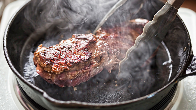 steak being cooked in a cast iron skillet with tongs and some smoke visible